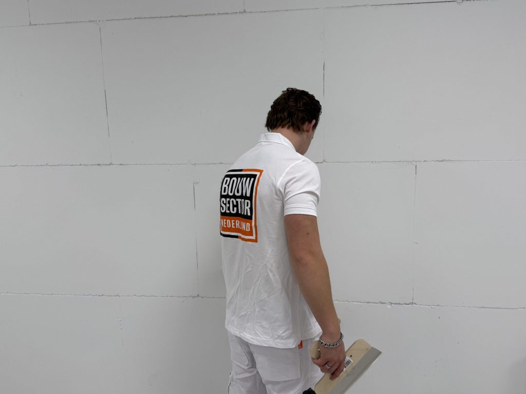 Stukadoor in wit poloshirt van Bouwsector Nederland staat met de spaan omlaag voor een wand van blokken en bekijkt de naden