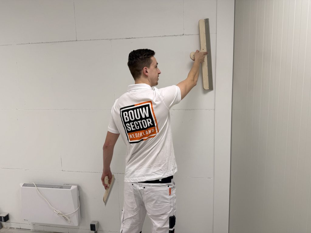 Stukadoor in witte werkbroek houdt een brede spaan tegen de bovenkant van de wand, kachel links onderin