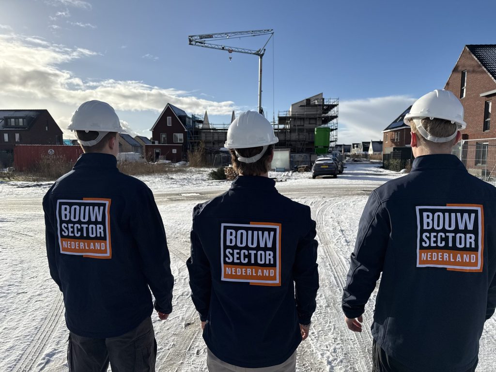 Drie bouwvakkers van Bouwsector Nederland kijken naar een rij nieuwbouwwoningen in aanbouw onder een blauwe winterlucht