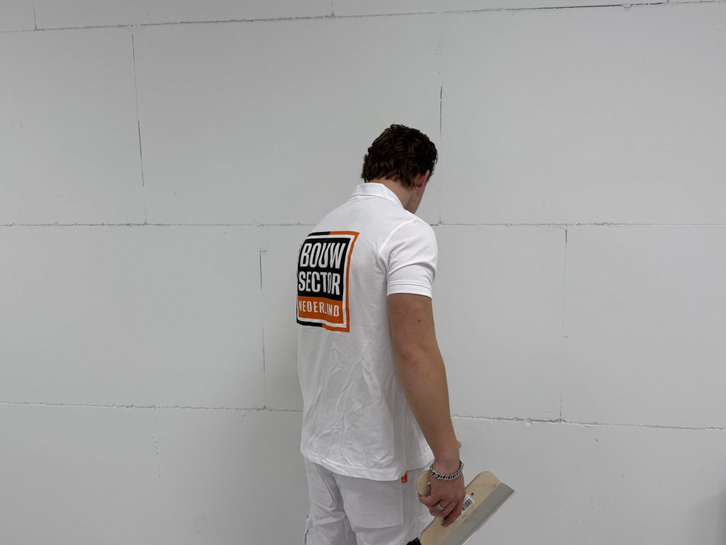 Stukadoor van Bouwsector Nederland in wit tenue voor een kale wand van gipsblokken, met een plakspaan in de hand